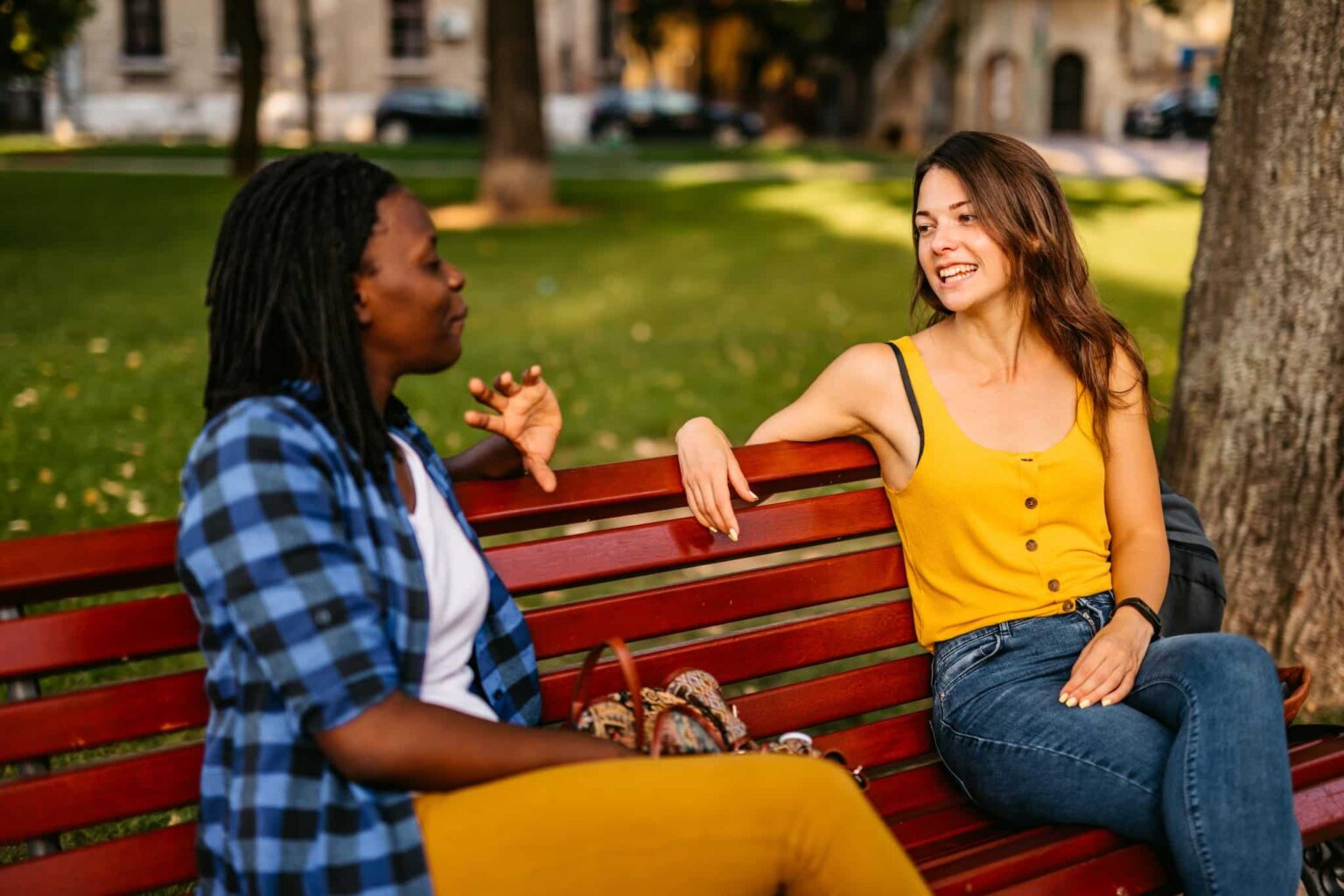 Two woman sit on a park bench having a discussion. The woman on the left is mid sentence and the woman on the right is watching her and smiling. 